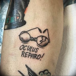 Черно-белое фото тату на руке с изображением сломанных очков и надписью 'Oculus Reparo!'. Стиль татуировки - минимализм, с четкими контурами и небольшим количеством деталей.