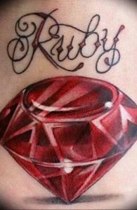 Фото тату рубина и надписи 'Ruby' выполненной каллиграфическим шрифтом. Яркий рисунок татуировки в красных и черных тонах.