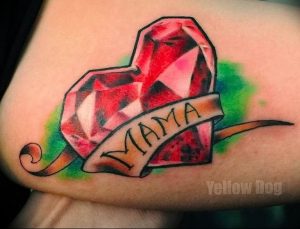 Фото тату на руке: красное сердце-рубин с надписью "MAMA" на ленте. Детализированный рисунок татуировки в стиле традишнл.