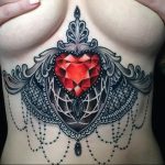Фото тату на груди, изображающее рубин, окруженный кружевным узором. Детализированный рисунок, черный и красный цвета, женственная татуировка.