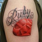 Фото тату рубина на плече с надписью 'Ruby'. Реалистичное изображение драгоценного камня с яркими красными оттенками.
