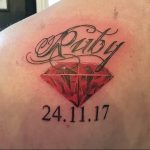 Фото тату рубина на плече с надписью 'Ruby' и датой '24.11.17'. Красный драгоценный камень реалистично изображен с бликами и тенями.