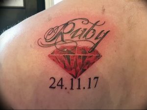 Фото тату рубина на плече с надписью 'Ruby' и датой '24.11.17'. Красный драгоценный камень реалистично изображен с бликами и тенями.
