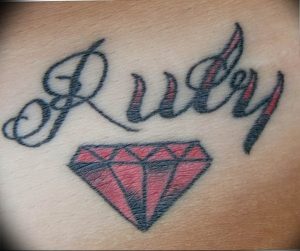 Фото тату с надписью 'Ruby' выполненной каллиграфическим шрифтом и красным бриллиантом под ней. Татуировка расположена на коже, демонстрируя изящный рисунок.