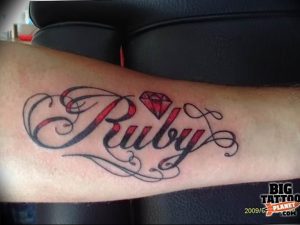 Фото тату на запястье с надписью 'Ruby' красивым шрифтом и небольшим красным бриллиантом внизу.