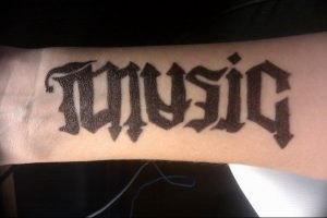 Фото тату с надписью "MUSIC" в готическом стиле шрифта. Черно-белый рисунок татуировки на запястье.