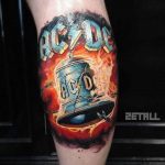 Фото тату на икре: красочный рисунок логотипа рок-группы AC/DC с изображением колокола и надписью, выполненный в стиле реализм с эффектом пламени.