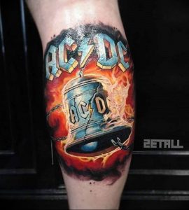 Фото тату на икре: красочный рисунок логотипа рок-группы AC/DC с изображением колокола и надписью, выполненный в стиле реализм с эффектом пламени.