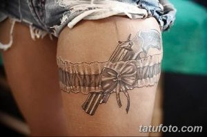 Фото тату пистолета, обвитого кружевной лентой с бантом на бедре. Черно-белый рисунок, сочетающий в себе брутальность и женственность.