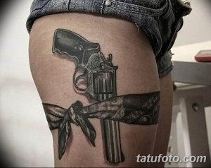 Фото тату револьвера с подвязкой на женском бедре. Рисунок татуировки в реалистичном стиле с использованием черных и серых оттенков.