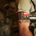 Фото тату на предплечье мужчины, изображающее башни-близнецы, горящие в огне, и надпись 'Can't Forget'. Рисунок татуировки выполнен в реалистичном стиле.