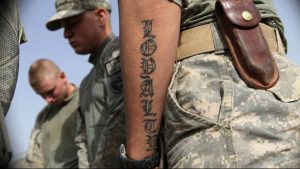 Фотография военного в камуфляжной форме с татуировкой надписью "LOYALTY" на руке. На заднем плане видны другие солдаты.
