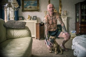 Фото пожилого мужчины с обширной татуировкой, покрывающей почти все тело. Он сидит с французским бульдогом.