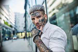 Фотография мужчины с множеством татуировок на лице, шее и руках. Мужчина одет в белую рубашку и выглядит уверенно.