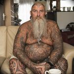Фотография пожилого мужчины с длинной седой бородой, чье тело полностью покрыто сложными татуировками в стиле трайбл и маори.