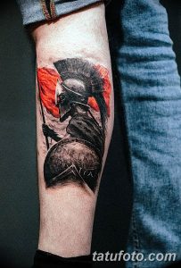 Фото тату на ноге: спартанский воин в шлеме и с щитом. Реалистичный рисунок татуировки в черно-серых тонах с элементами красного.