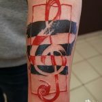 Фото абстрактной татуировки с изображением скрипичного ключа на руке, выполненное в красных, черных и белых тонах. Рисунок татуировки с геометрическими элементами.