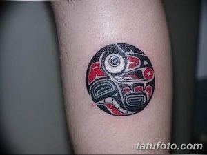 Фото тату в стиле Northwest Coast Native на руке. Круглый рисунок с изображением головы животного, выполненный в черном и красном цветах.