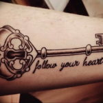 Фото тату: реалистичный ключ с витиеватой головкой и надписью 'Follow Your Heart' на предплечье. Черно-серый рисунок татуировки.
