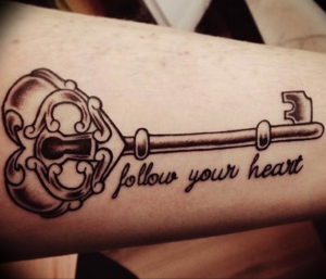 Фото тату: реалистичный ключ с витиеватой головкой и надписью 'Follow Your Heart' на предплечье. Черно-серый рисунок татуировки.