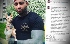 Фотография мускулистого мужчины с татуировкой на плече и груди, обнимающего белую кошку. Мужчина в спортивной одежде, на фоне комната.