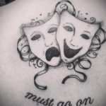 Фото тату на груди: театральные маски трагедии и комедии с надписью "The show must go on", выполненное в технике графики.