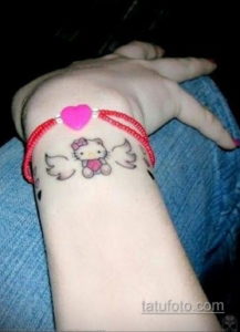 Фото тату Hello Kitty с крыльями и сердцем на запястье. Черно-белый рисунок с розовым сердцем символизирует нежность и любовь.