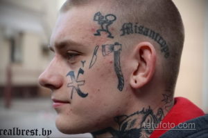 Фото мужчины с множеством татуировок на лице, шее и голове. Различные символы, надписи и рисунки, выполненные черным цветом. Экстремальный и вызывающий образ.