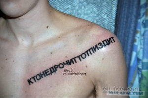 Фото татуировки с неразборчивой надписью на груди мужчины. Рисунок татуировки выполнен черными чернилами.