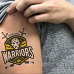 Фото тату с изображением логотипа хоккейной команды 'Warriors' на плече мужчины. Черно-белый рисунок татуировки в стиле олдскул с элементами экипировки.