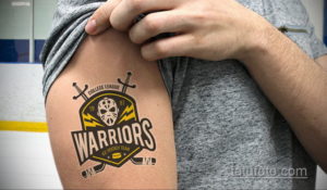 Фото тату с изображением логотипа хоккейной команды 'Warriors' на плече мужчины. Черно-белый рисунок татуировки в стиле олдскул с элементами экипировки.