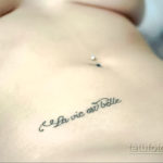 Фото тату на боку: изящная надпись "La vie est belle" (Жизнь прекрасна) каллиграфическим шрифтом. Минималистичная и вдохновляющая татуировка.