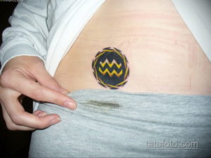 Фото тату: Цветная татуировка символа Водолея на женском животе. Символ заключен в круг с декоративными элементами.
