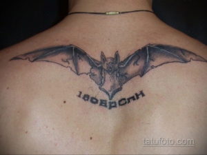 Фото тату летучей мыши на спине мужчины. Черно-серый рисунок с надписью, выполненный в реалистичном стиле. Идеально для любителей готики и мистики.