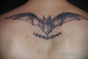 Фото черно-серой татуировки летучей мыши на спине мужчины. Детализированный рисунок с надписью под крыльями.