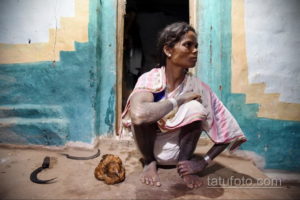 Фотография индийской женщины с традиционными татуировками на руках и ногах, сидящей у входа в дом.