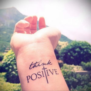 Фото тату: Надпись "Think Positive" с изображением креста на запястье. Татуировка с позитивным посланием и религиозным символом.