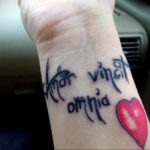 Фото тату: надпись "Amor vincit omnia" и красное сердце на запястье. Черные линии, каллиграфический шрифт, романтическое значение.