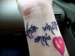 Фото тату: надпись "Amor vincit omnia" и красное сердце на запястье. Черные линии, каллиграфический шрифт, романтическое значение.