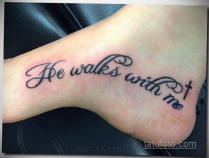 Фото тату с надписью 'He walks with me' и небольшим крестом на стопе. Черно-белая татуировка, выполненная аккуратным шрифтом.