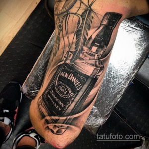 Фото тату реалистичной бутылки виски Jack Daniel's и этикетки на руке мужчины, выполненное в черно-серых тонах с детальной проработкой.