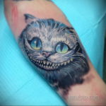 Фото реалистичной татуировки Чеширского кота из Алисы в Стране чудес на руке. Кот с широкой улыбкой и яркими глазами.