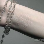 Фото белой татуировки снежинки на предплечье. Детализированный рисунок с тонкими линиями, создающий эффект легкости и воздушности. На фоне бусы.