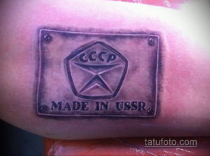 Фото тату на руке с изображением советского герба, стилизованного под значок 'Made in USSR', выполненное в черно-серых тонах.