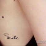 Фото тату с надписью "Smile" на груди. Рисунок выполнен в черном цвете, каллиграфическим шрифтом.