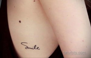 Фото тату с надписью "Smile" на груди. Рисунок выполнен в черном цвете, каллиграфическим шрифтом.