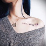 Фото тату: вдохновляющая надпись 'Love thyself' (люби себя) выполнена каллиграфическим шрифтом и расположена на ключице девушки.