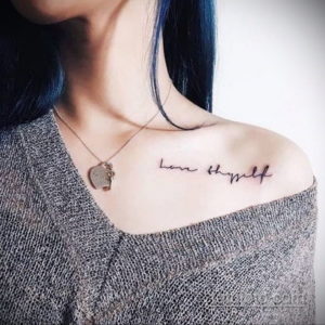 Фото тату: вдохновляющая надпись 'Love thyself' (люби себя) выполнена каллиграфическим шрифтом и расположена на ключице девушки.