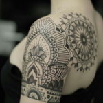 Фото тату: сложная мандала, выполненная в технике dotwork, покрывает плечо и часть руки девушки. Черно-белая татуировка с геометрическими узорами.