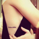 Фото тату: надпись "Live a little" на плече девушки. Небольшая, но вдохновляющая татуировка, выполненная черными чернилами.